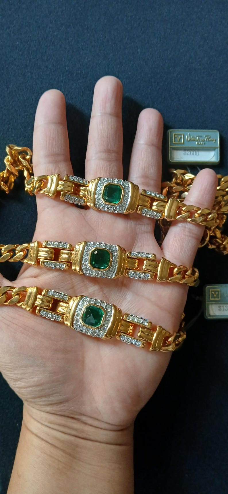 May include: Gold-toned bracelets with emerald-colored center stones and clear accent stones. The bracelets are displayed on a hand, with a gold chain visible. The bracelets have a rectangular design.