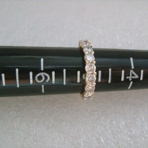 May include: A gold band with a row of small, clear round stones. The band is on a ruler, measuring approximately 1.25 inches in diameter.