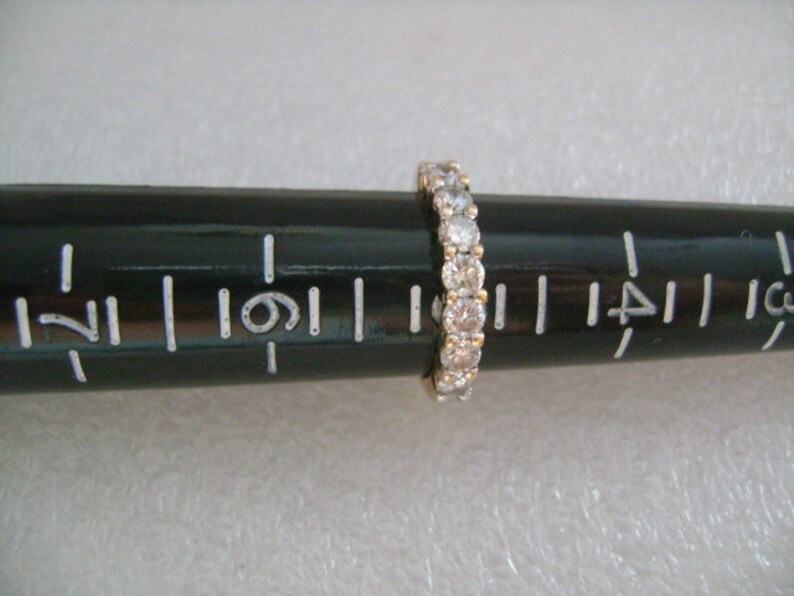 May include: A gold band with a row of small, clear round stones. The band is on a ruler, measuring approximately 1.25 inches in diameter.
