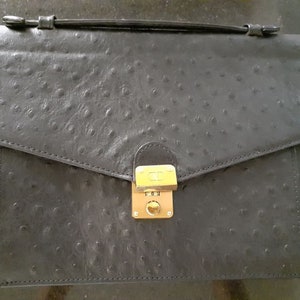 May include: A black ostrich leather briefcase with a gold lock and clasp. The briefcase has a flap that closes with a buckle.