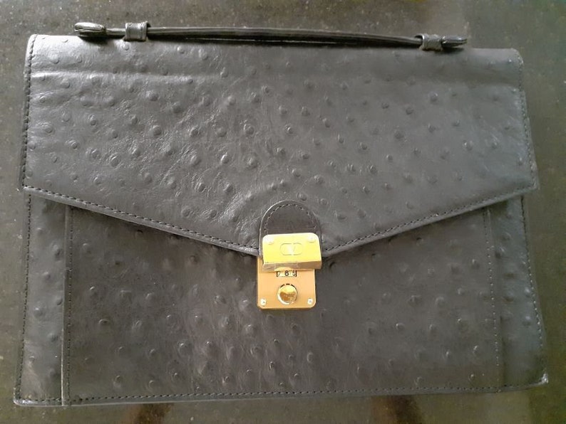 May include: A black ostrich leather briefcase with a gold lock and clasp. The briefcase has a flap that closes with a buckle.