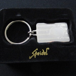 May include: A silver-colored keychain with a rectangular tag featuring an oval design and horizontal lines, presented in a black velvet box. The keychain has a circular ring and the brand name "Speidel" in gold lettering.