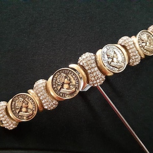 May include: A gold bracelet with a series of round, coin-shaped charms featuring a profile of a woman's face. The charms are surrounded by clear rhinestones.
