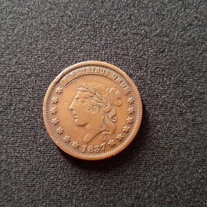 May include: A copper-colored coin with the inscription "United States of America" and the year "1857" on the front. The coin features a portrait of a woman with her hair pulled back.