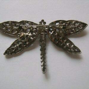 May include: Silver-toned dragonfly brooch with intricate cut-out details. The brooch has a pin and catch closure on the back.