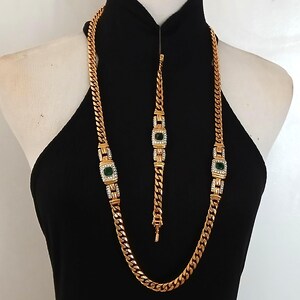 May include: Gold-toned necklace and bracelet set. The necklace and bracelet feature a curb chain design with square emerald-green and clear crystal accents. The jewelry is displayed on a black dress form.
