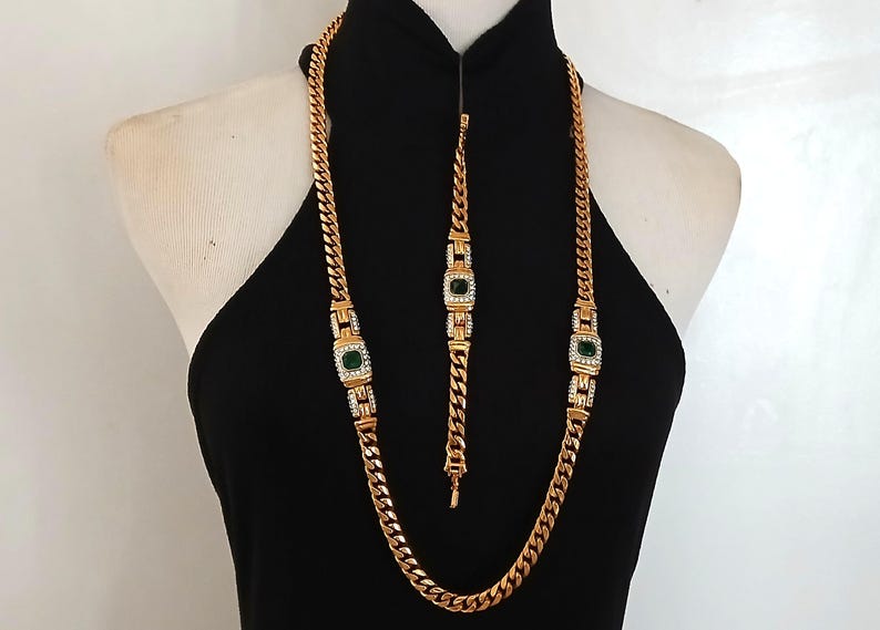 May include: Gold-toned necklace and bracelet set. The necklace and bracelet feature a curb chain design with square emerald-green and clear crystal accents. The jewelry is displayed on a black dress form.