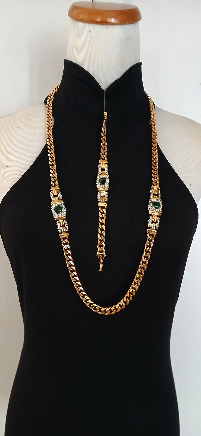 May include: Gold-toned necklace and bracelet set. The necklace and bracelet feature a curb chain design with emerald-green and clear crystal accents. The jewelry is displayed on a black mannequin.