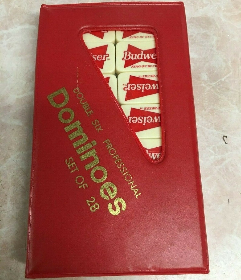 May include: A red, rectangular box with gold lettering that reads "Dominoes Double Six Professional Set of 28". The box has a clear plastic window that shows a set of dominoes with the Budweiser logo.