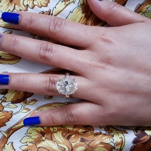 Op de afbeelding: Een grote, ronde, heldere diamanten ring gezet in een zilveren band. De ring zit aan een vinger met blauwe nagellak.