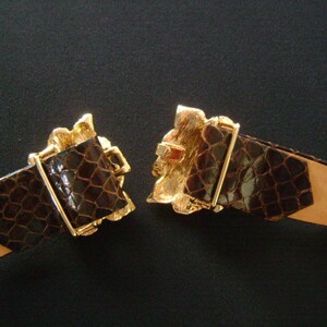 May include: A brown leather belt with a gold-tone buckle featuring an ornate design. The buckle is open and the belt is lying flat.