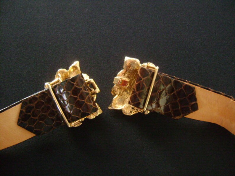 May include: A brown leather belt with a gold-tone buckle featuring an ornate design. The buckle is open and the belt is lying flat.