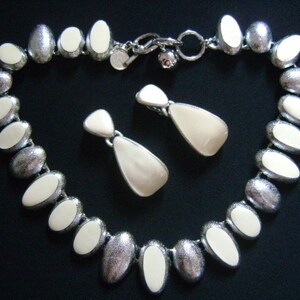 Puede incluir: Un collar y pendientes a juego de forma ovalada plateados y blancos. El collar tiene una cadena plateada con cuentas ovaladas blancas. Los pendientes son plateados con piedras ovaladas blancas.