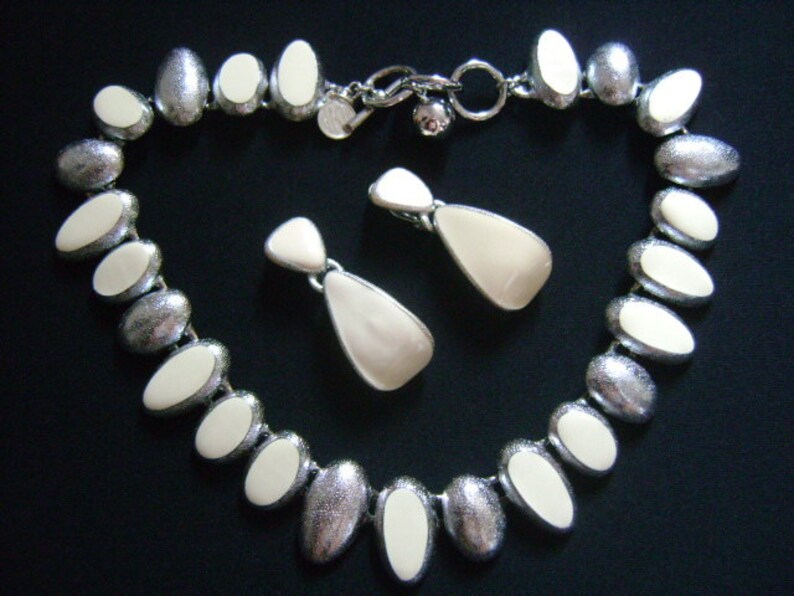 Puede incluir: Un collar y pendientes a juego de forma ovalada plateados y blancos. El collar tiene una cadena plateada con cuentas ovaladas blancas. Los pendientes son plateados con piedras ovaladas blancas.