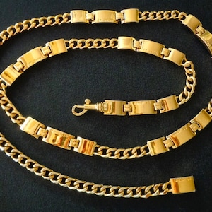 May include: A gold chain necklace with a thick, flat link design. The chain is made of a series of connected rectangular links.