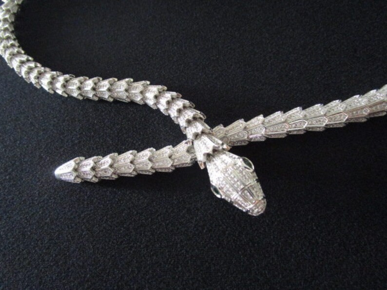 Op de afbeelding: Een zilveren slangenketting met een diamantbezette ontwerp. De kop van de slang is naar voren gericht met de mond open.