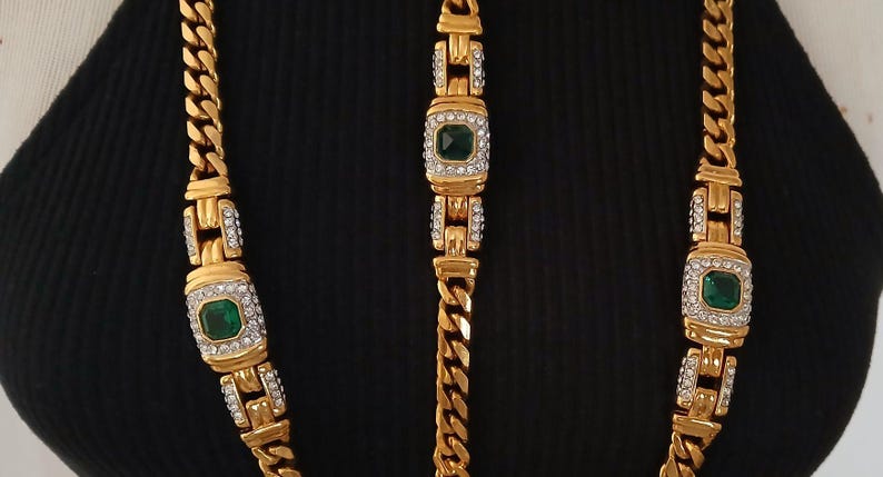 May include: Gold-toned necklace and bracelet set. The necklace and bracelet feature a curb chain design with rectangular links. Each link has a square emerald-green stone surrounded by clear crystals.