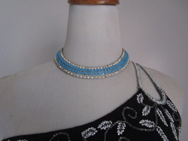 Puede incluir: Un collar gargantilla de pedrer&iacute;a azul y plata en un maniqu&iacute; blanco. El collar tiene tres filas de pedrer&iacute;a cuadrada.