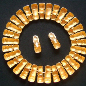 May include: A gold-tone necklace with a chain made of linked rectangular pieces. The necklace is adorned with small, clear rhinestones. The necklace is displayed on a black surface with a pair of matching earrings.