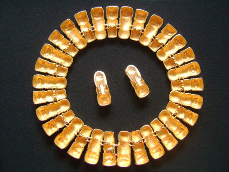 May include: A gold-tone necklace with a chain made of linked rectangular pieces. The necklace is adorned with small, clear rhinestones. The necklace is displayed on a black surface with a pair of matching earrings.