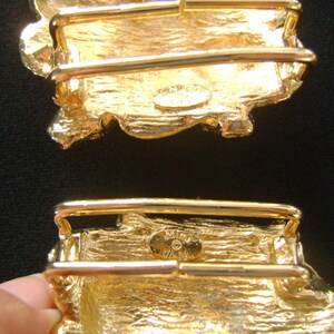 May include: Two gold-tone belt buckles with a textured, irregular shape. The buckles have a rectangular design with a raised, textured surface. The buckles are designed to be used with a belt.