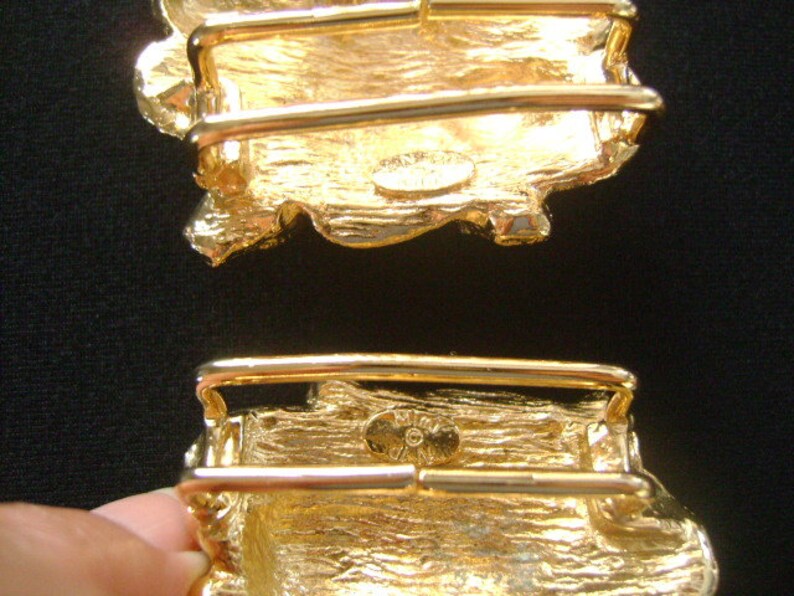 May include: Two gold-tone belt buckles with a textured, irregular shape. The buckles have a rectangular design with a raised, textured surface. The buckles are designed to be used with a belt.