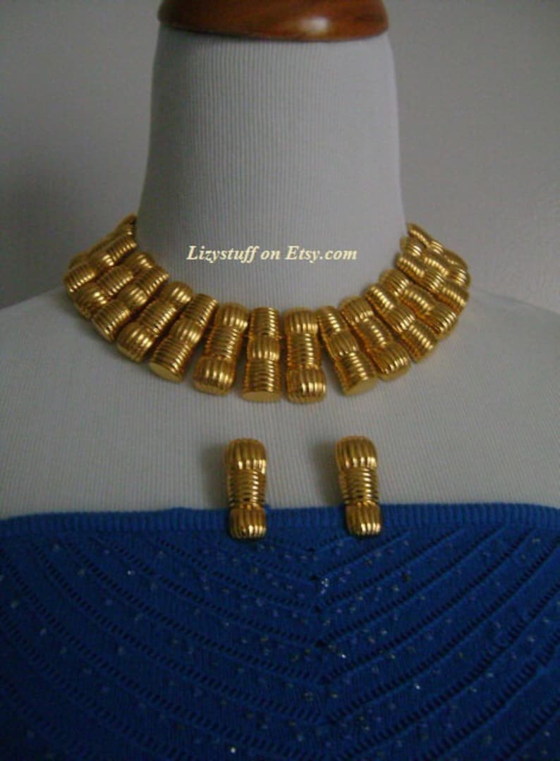 May include: A gold-tone statement necklace and matching earrings. The necklace is made of a series of linked, ridged, gold-tone cylinders. The earrings are simple, gold-tone cylinders.  Lizystuff on Etsy.com