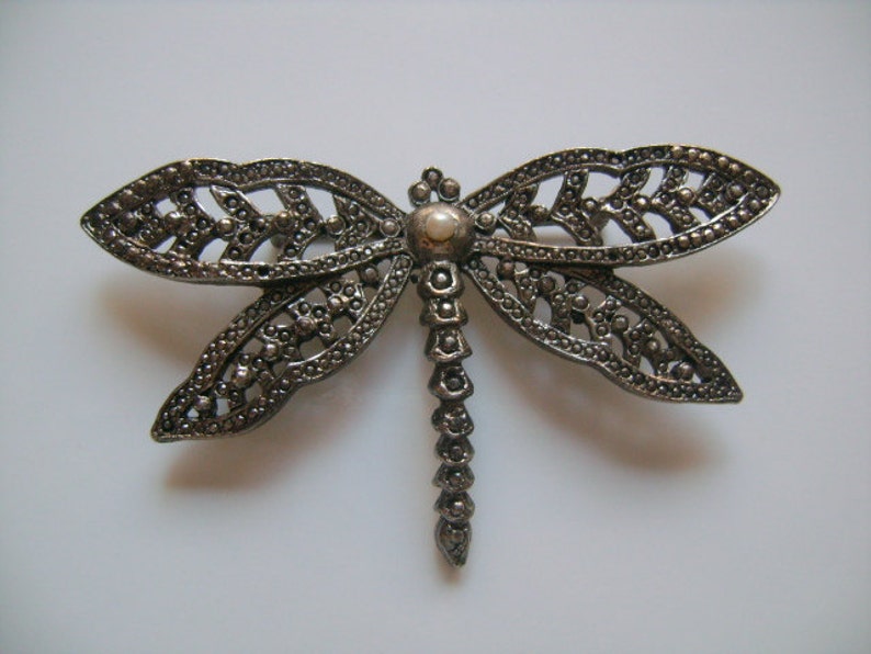May include: A silver dragonfly brooch with a pearl-like center. The wings are detailed with intricate filigree work.
