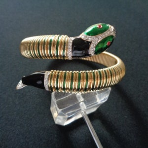 Könnte beinhalten: Ein goldfarbenes, grünes und schwarzes Schlangenarmband mit einem gewundenen Design und klaren Strasssteinen. Der Kopf der Schlange ist erhoben und hat rote Augen.