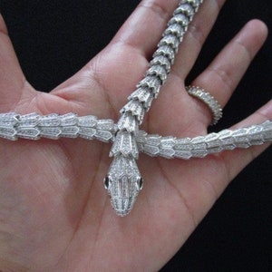 Op de afbeelding: Een zilveren slangenketting met een diamant-achtige afwerking. De ketting is in de vorm van een slang met zijn kop omhoog en zijn staart om de nek gewikkeld.