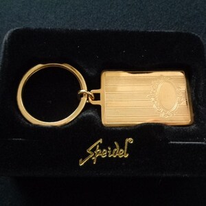 May include: Gold-coloured key chain with a rectangular tag that has an engraved design and the word "Speidel". The key chain is in a black velvet box.