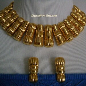 May include: A gold-tone statement necklace and matching earrings. The necklace is made of a series of linked, textured, gold-tone cylinders. The earrings are simple, single cylinder designs.