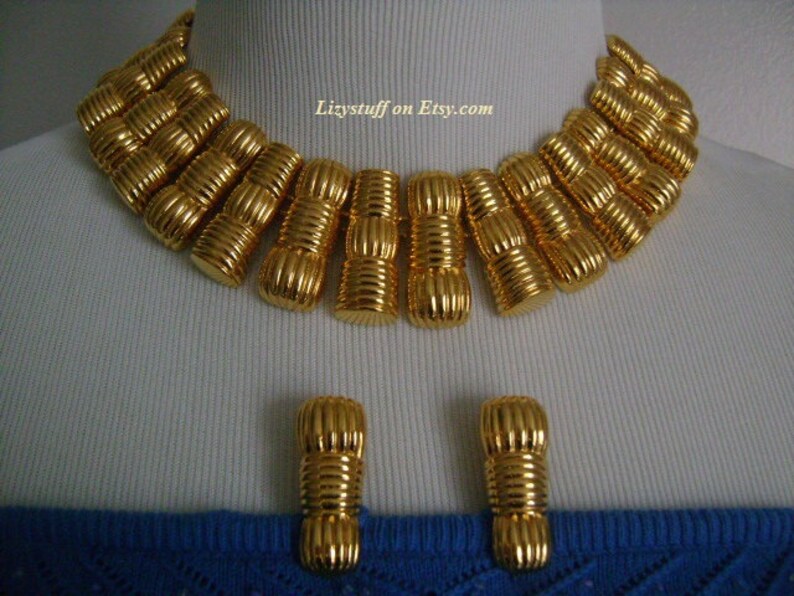 May include: A gold-tone statement necklace and matching earrings. The necklace is made of a series of linked, textured, gold-tone cylinders. The earrings are simple, single cylinder designs.
