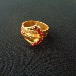 May include: A gold ring featuring a red stone design. The ring has a distinctive, elaborate design with multiple bands and a large red stone at the centre.