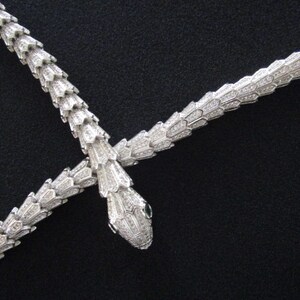 Op de afbeelding: Een zilveren slangenketting met een dubbel slangenontwerp. De ketting is gemaakt van metaal en heeft een glanzende afwerking. De slangen zijn met elkaar verweven en hebben een gedetailleerd ontwerp.