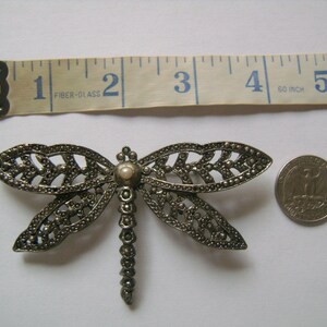 May include: A silver-toned dragonfly brooch with a pearl-like center. The brooch is about 2 inches wide and is shown next to a US quarter for size comparison.