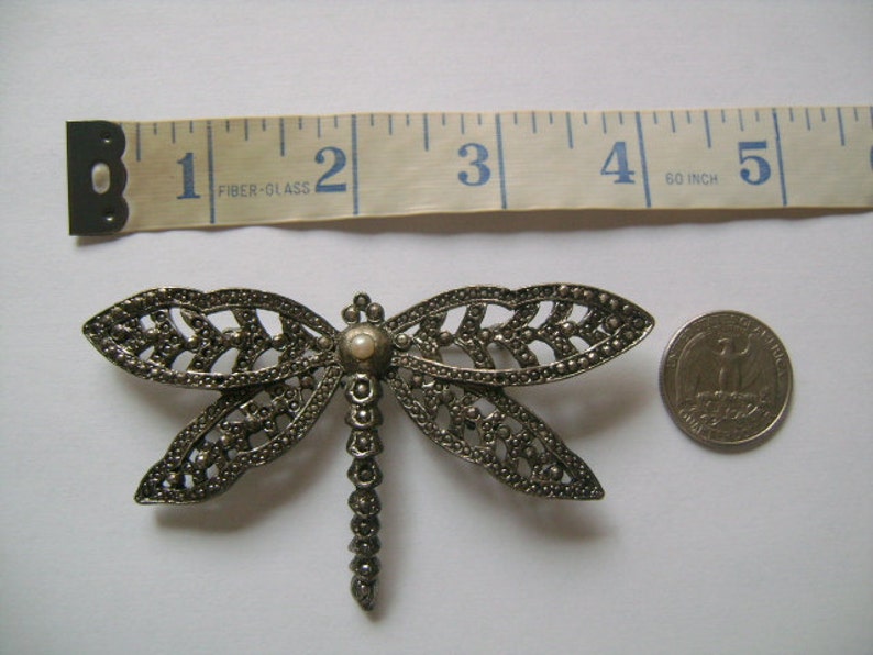 May include: A silver-toned dragonfly brooch with a pearl-like center. The brooch is about 2 inches wide and is shown next to a US quarter for size comparison.