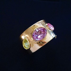May include: A gold bangle bracelet with three colorful gemstones. The bracelet has a wide band and features a large oval purple stone, a smaller oval pink stone, and a teardrop-shaped green stone.