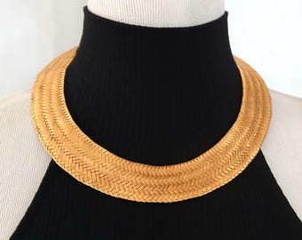 Puede incluir: Un collar tejido dorado. El collar es grueso y se asienta cerca del cuello, con un patrón texturizado en forma de espiga. El collar se exhibe sobre un top negro de cuello alto contra un fondo blanco.