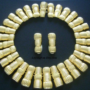 May include: A gold-tone necklace and earrings set with a geometric design. The necklace is made of a series of linked, textured, cylindrical pieces. The earrings are a matching pair of small, cylindrical pieces.