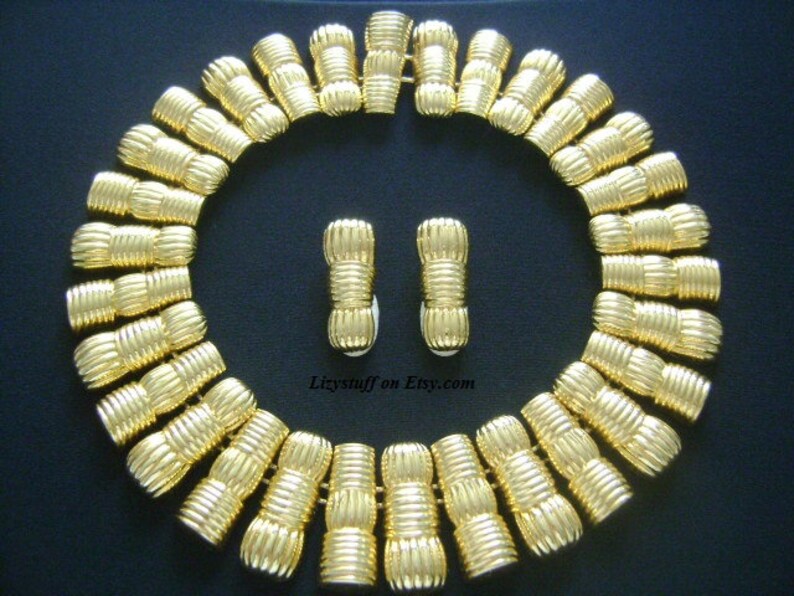 May include: A gold-tone necklace and earrings set with a geometric design. The necklace is made of a series of linked, textured, cylindrical pieces. The earrings are a matching pair of small, cylindrical pieces.