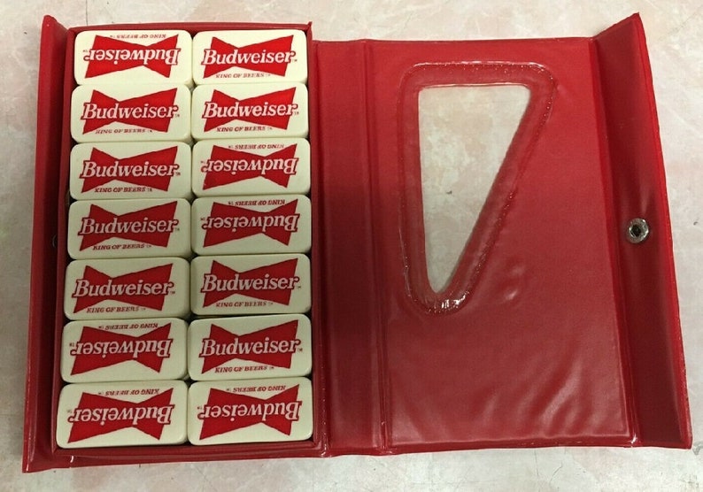 May include: A set of white dominoes with red Budweiser logos and the text "King of Beers". The dominoes are in a red case with a clear plastic window.
