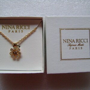 May include: A gold necklace with a flower-shaped pendant set with clear crystals. The necklace comes in a white box with the words "Nina Ricci Bijoux Mode Paris" printed on it.