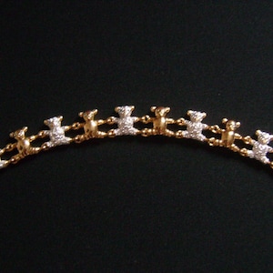 Op de afbeelding: Een gouden armband met een reeks kleine teddybeerfiguren. Sommige beren zijn versierd met heldere kristallen. De armband is geplaatst tegen een donkere achtergrond, waardoor het ontwerp wordt benadrukt.