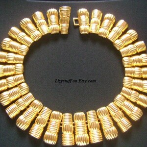 May include: A gold-tone necklace with a series of linked, ridged, cylindrical beads. The necklace is laid out on a black surface.