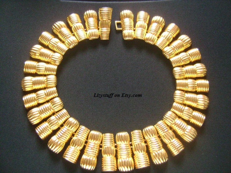 May include: A gold-tone necklace with a series of linked, ridged, cylindrical beads. The necklace is laid out on a black surface.