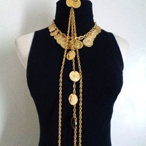 May include: A gold necklace with multiple strands of chain and coin-shaped charms. The necklace is long and hangs down to the chest. The necklace is worn on a black mock neck shirt.