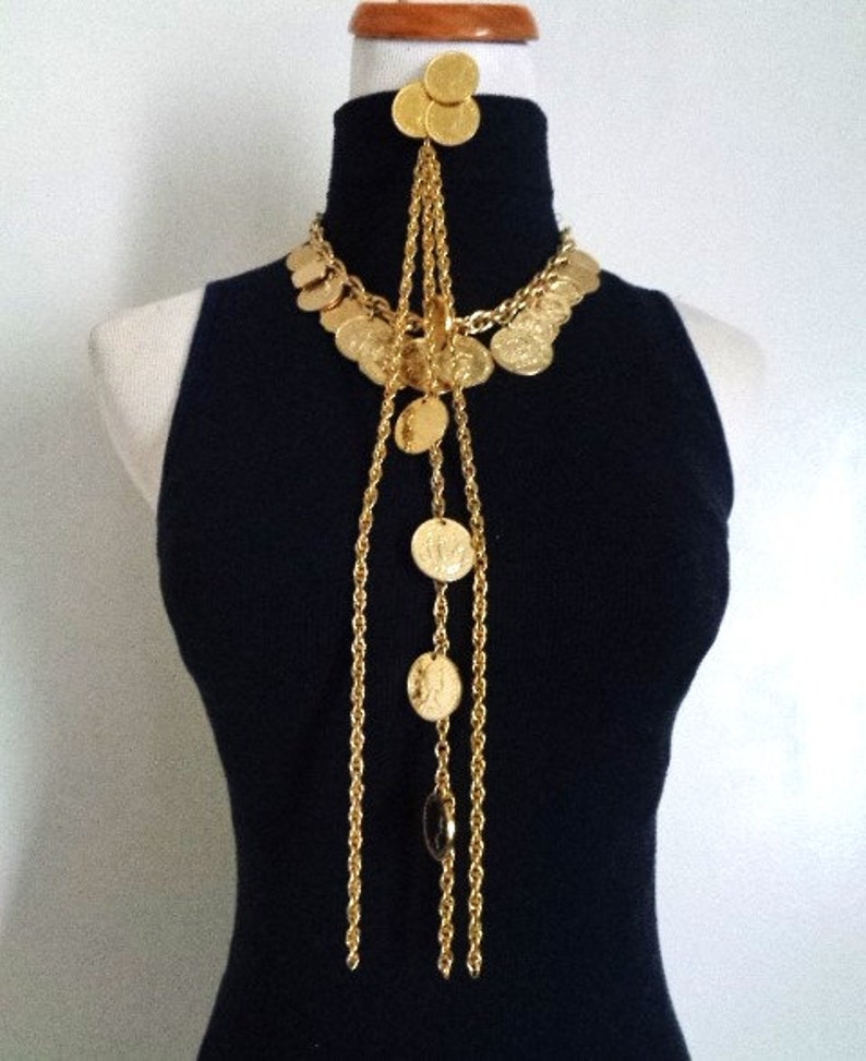 May include: A gold necklace with multiple strands of chain and coin-shaped charms. The necklace is long and hangs down to the chest. The necklace is worn on a black mock neck shirt.