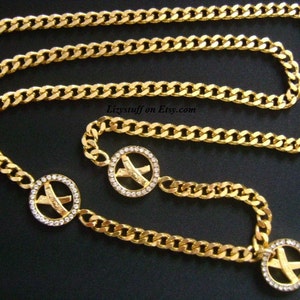 Puede incluir: Un cinturón de cadena dorada con tres colgantes circulares. Cada colgante presenta un diseño en X dorado con diamantes de imitación transparentes.