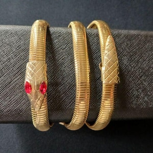 May include: Gold-toned snake bracelet with red gemstone eyes. The snake's head is detailed with a textured pattern, and the body coils around itself. The bracelet is displayed on a dark gray surface, showcasing its intricate design.